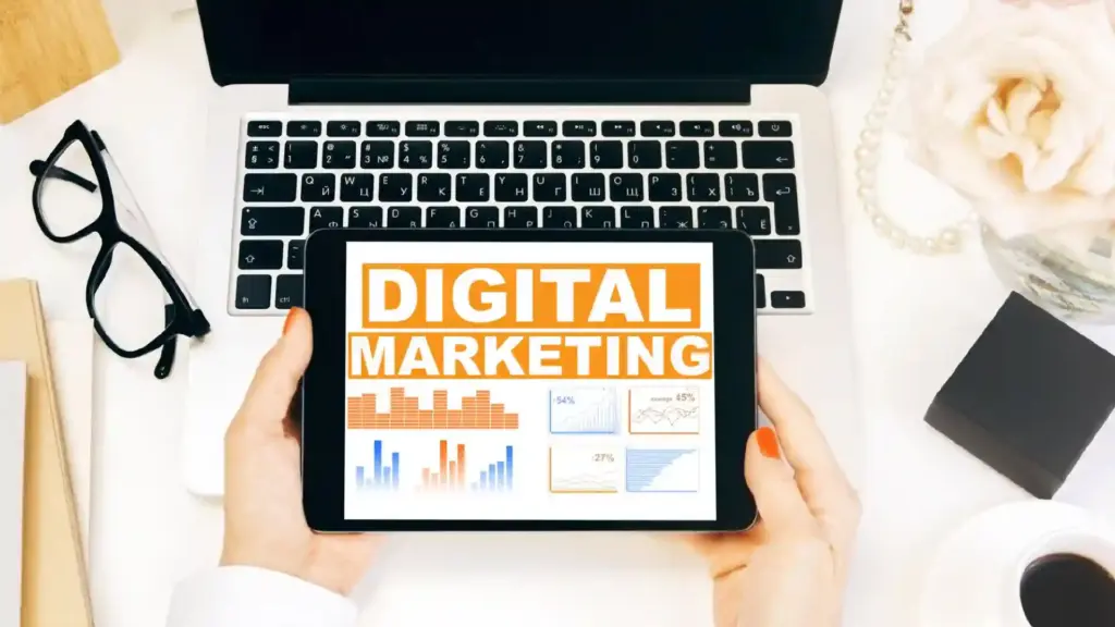 Tablet displaying 'DIGITAL MARKETING' graphics and charts held over a laptop keyboard on a desk.