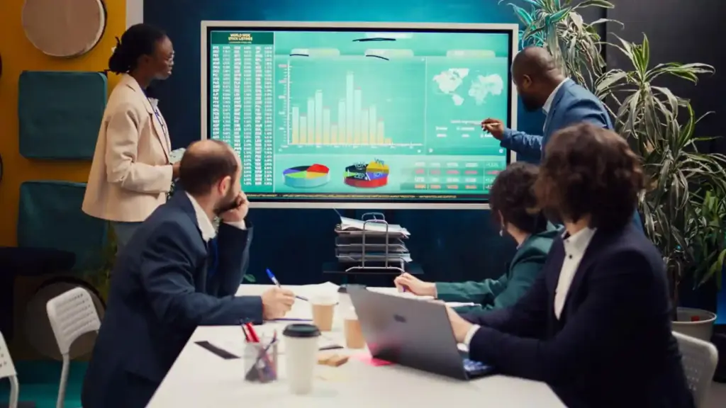 Team in a conference room reviews data on a large touchscreen board with charts; a man points to the screen while colleagues take notes.