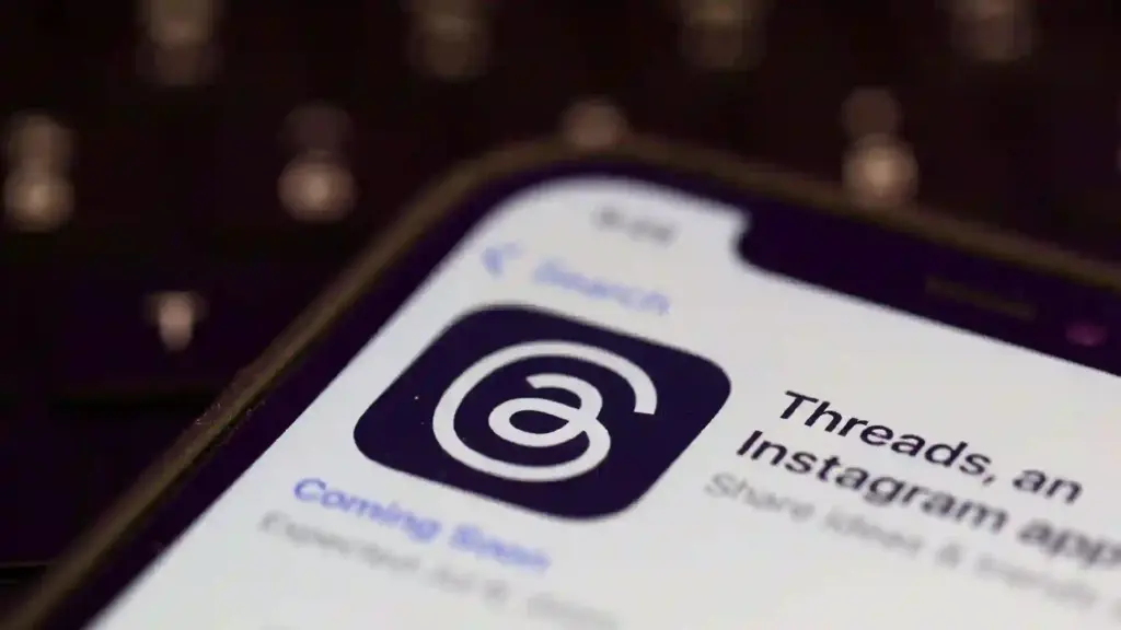 Close-up of a smartphone screen displaying an app icon in a dark rounded square with a white spiral, with blurred city lights in the background (likely Threads by Instagram).