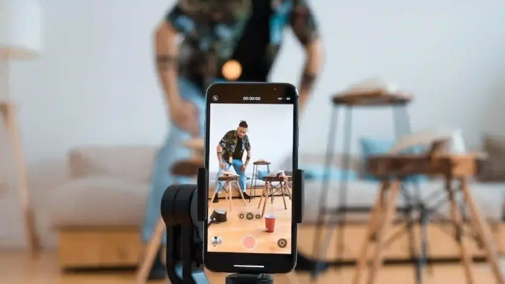 Person filming with a smartphone mounted on a tripod in a bright living room, with a man assembling small stools in the background on the phone screen.