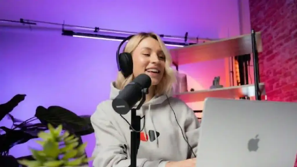 Person wearing headphones speaks into a microphone in a podcast studio, with a laptop in front and purple lighting in the background.