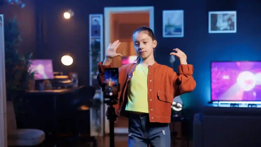 Young girl filming herself in a living room with a smartphone on a tripod, posing with hands up.