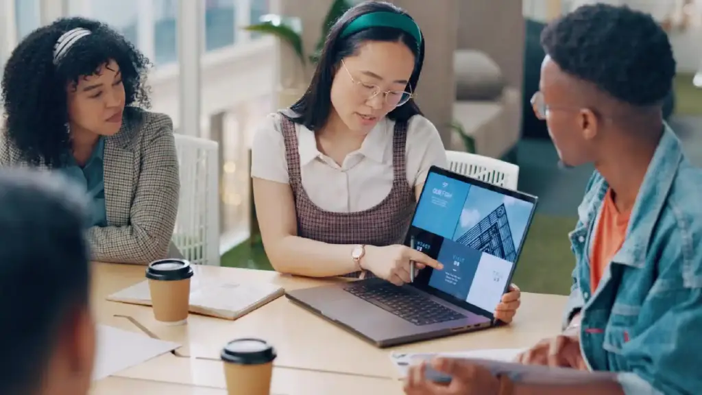 Group of coworkers in a meeting around a table; a woman points at a laptop screen displaying a blue architectural slide.