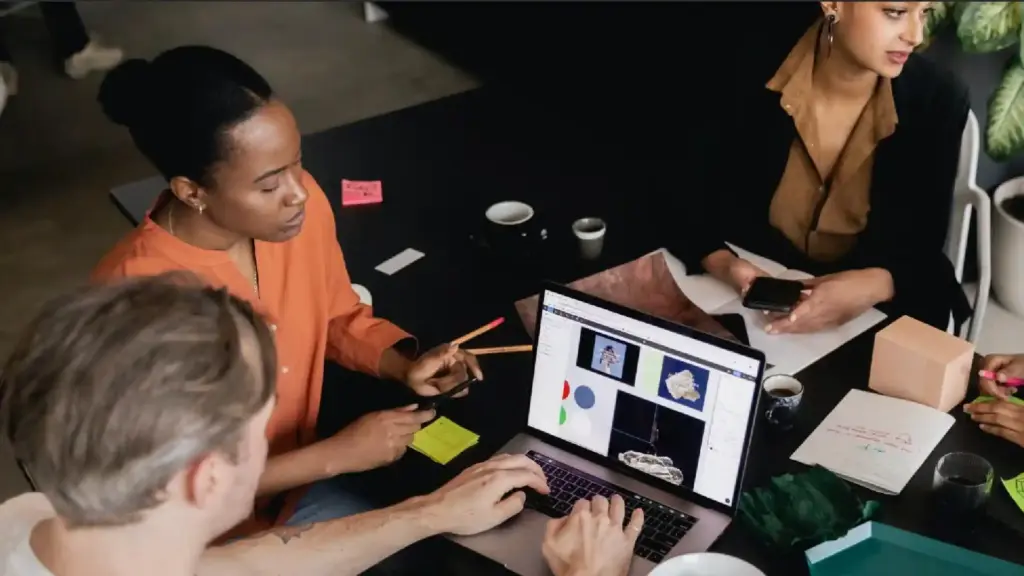 A group of people collaborating around a table, looking at a laptop showing design images and charts, with notes and cups on the table.