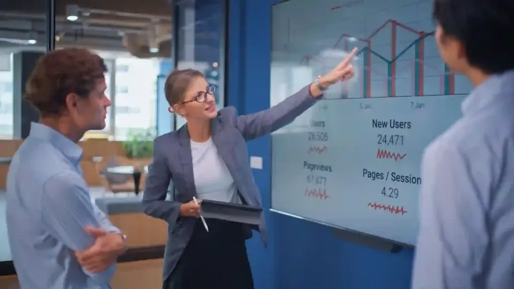 New Project - 2026-06-01T131652947 - Brill Creations Businesswoman points to a large wall screen showing charts as two colleagues watch in a modern office conference area.