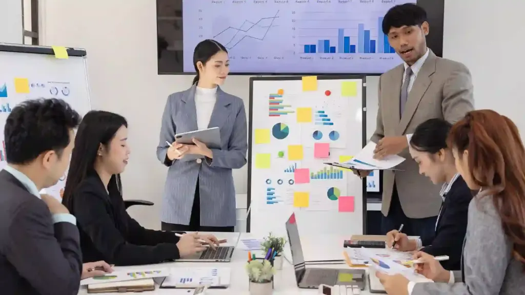 Presenter explains data to a group around a conference table; colorful charts and sticky notes on a whiteboard behind them.