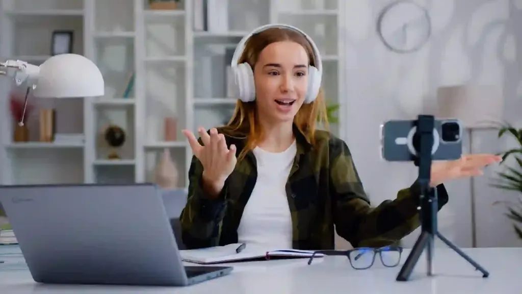 Woman wearing white over-ear headphones speaks to a camera while gesturing at a desk setup for a video recording or livestream.