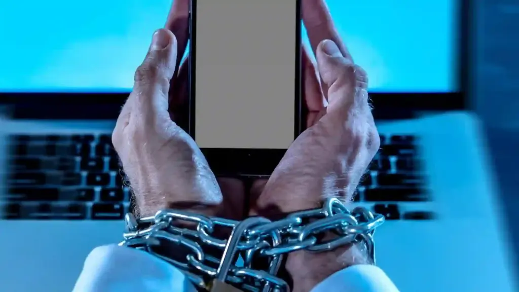 Hands bound with a metal chain grip a smartphone with a blank screen; a laptop keyboard appears in the blue background.
