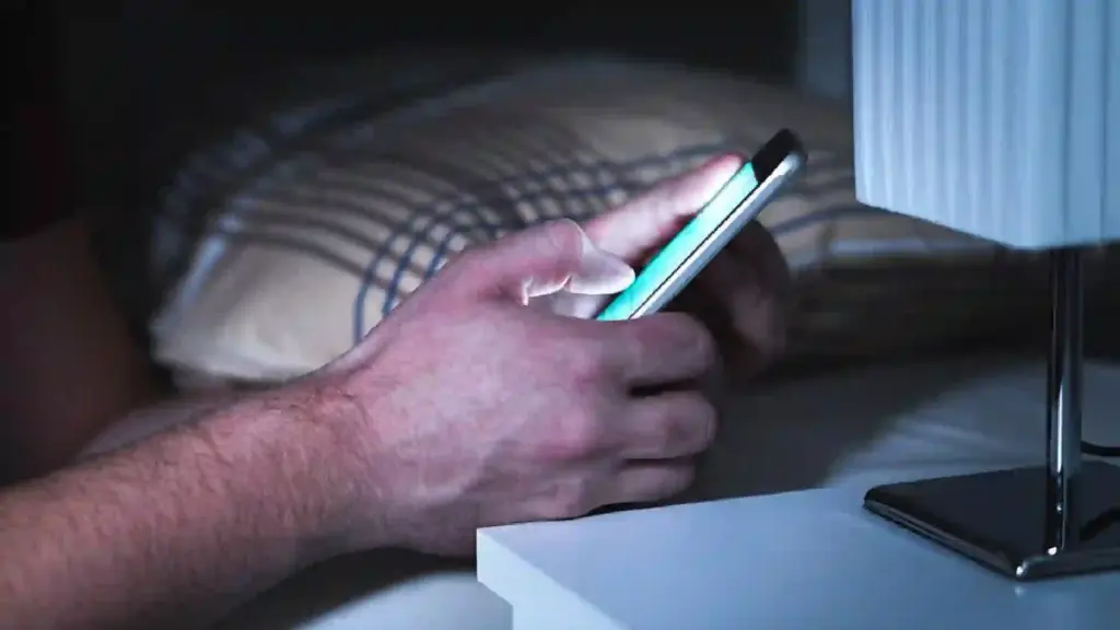 Close-up of a hand holding a smartphone with a bright screen on a white desk in low light.