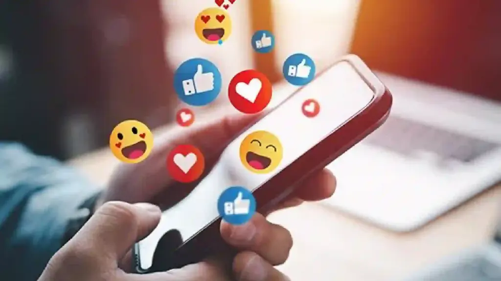 Person holds a smartphone with floating social media reaction icons (like, heart, emoji) around the screen.
