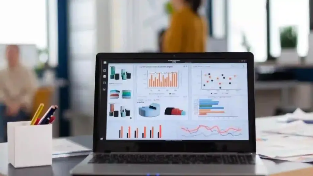 Laptop on a desk showing a multi-chart analytics dashboard; blurred office background with a person in orange.