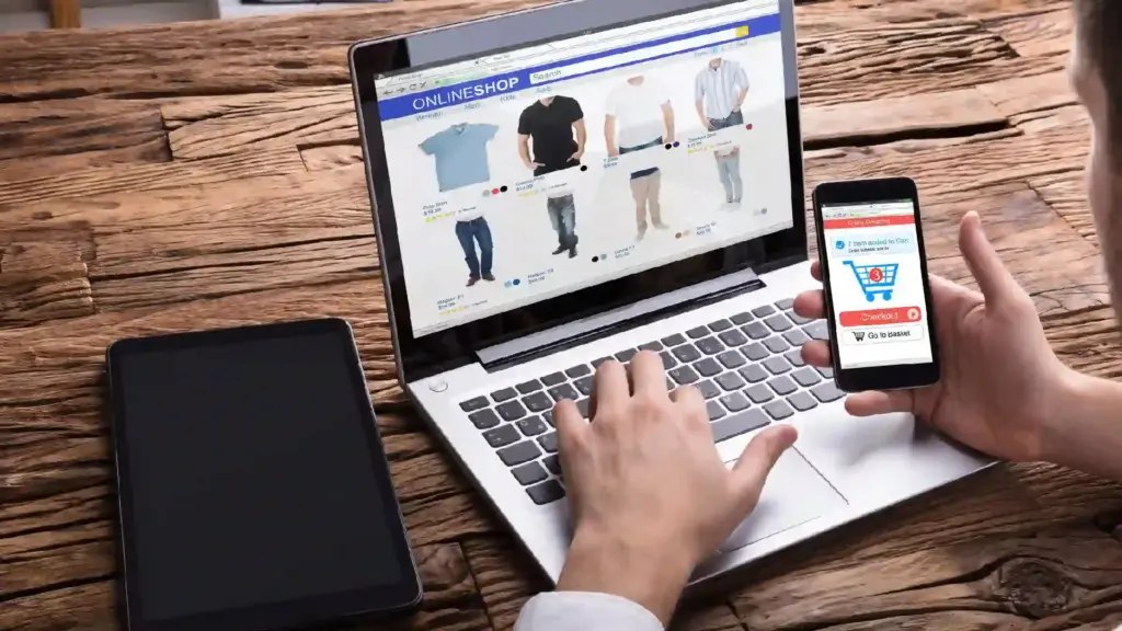 New Project - 2026-06-21T120020921 - Brill Creations Person browses an online clothing store on a laptop while holding a smartphone displaying a checkout screen