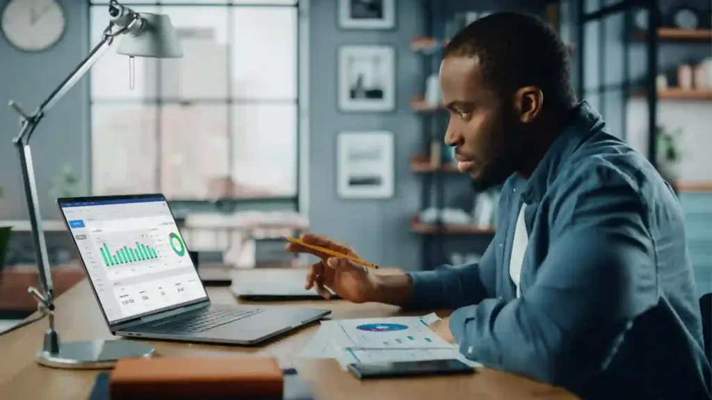 New Project - 2026-06-21T120327963 - Brill Creations Man analyzing data on a laptop while holding a pencil at a cluttered desk with charts on the screen and documents nearby in a modern office.