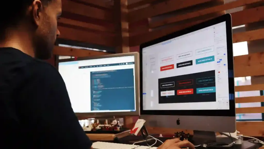 Person seated at a desk using two large monitors, one showing code and the other a design/dashboard interface.