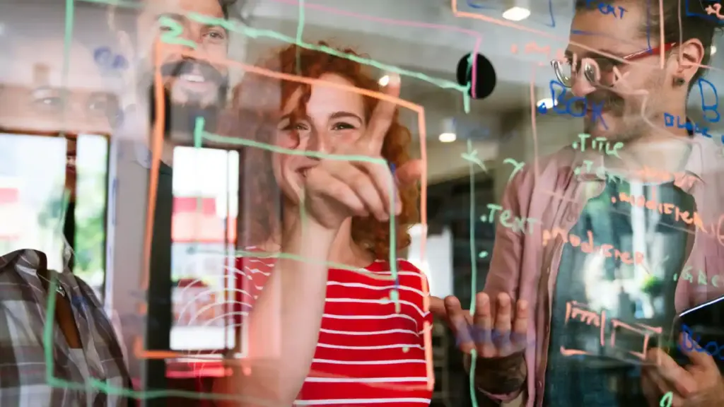 Two coworkers stand behind a glass board covered in colorful marker notes, smiling at the camera; woman in a red striped shirt in the center, man with glasses on the right.