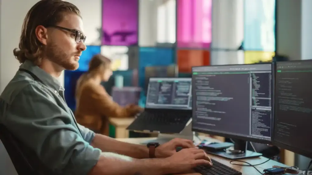 Profile of a man with glasses coding at a desk with multiple monitors displaying code in a bright office.