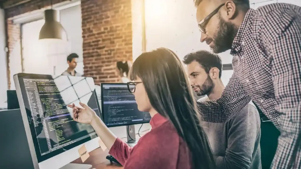 Team of developers collaborating at shared desks, reviewing code on large monitor screens in a brick-walled office.