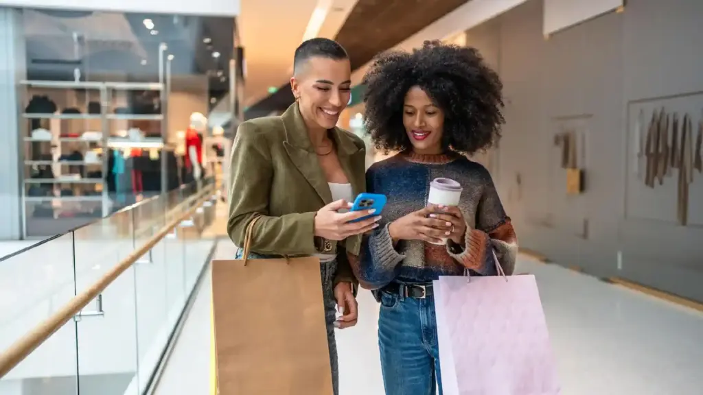 Two friends shopping in a mall, smiling and looking at a smartphone while holding shopping bags and a coffee cup.