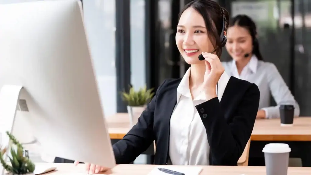 Smiling woman in a blazer wearing a headset and working at a computer in an office setting.