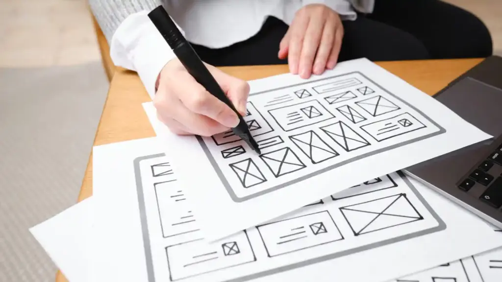 Person sketching website wireframes on paper with a marker, laptop nearby on the desk