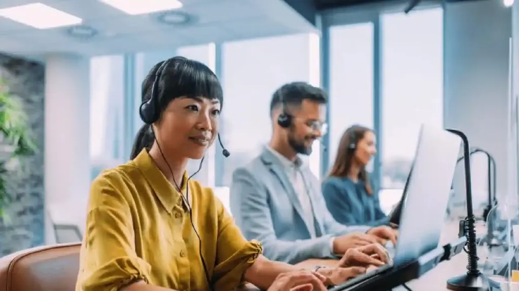 Customer service representative wearing a headset and typing at a computer in a bright office.