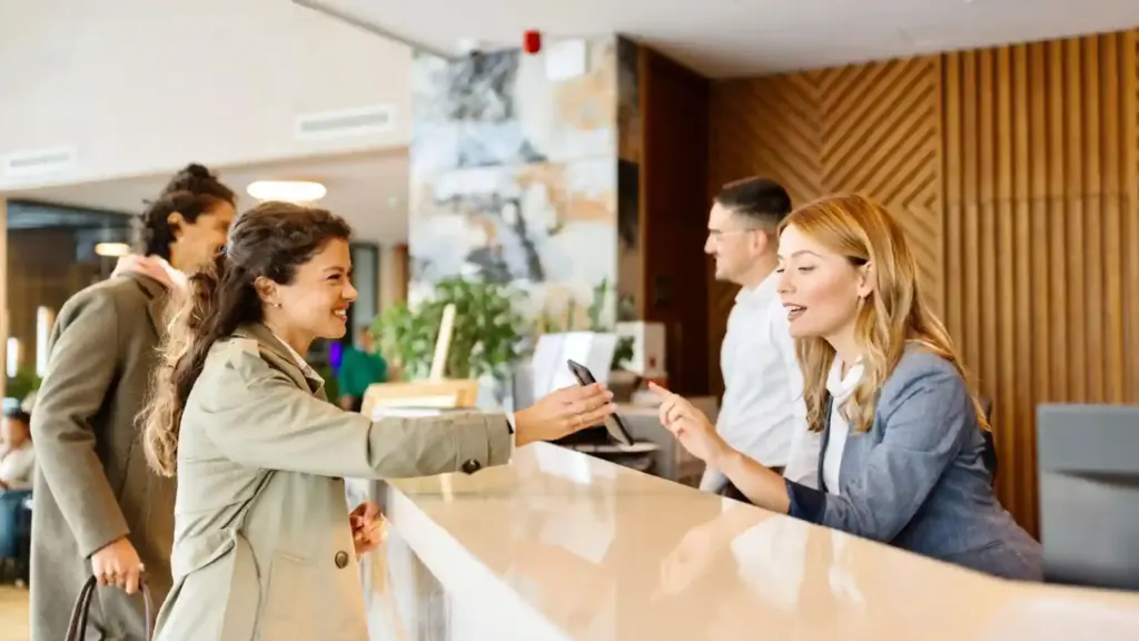 Guest handover at a bright hotel front desk; staff member accepts a phone with a smile in a modern lobby reception area.