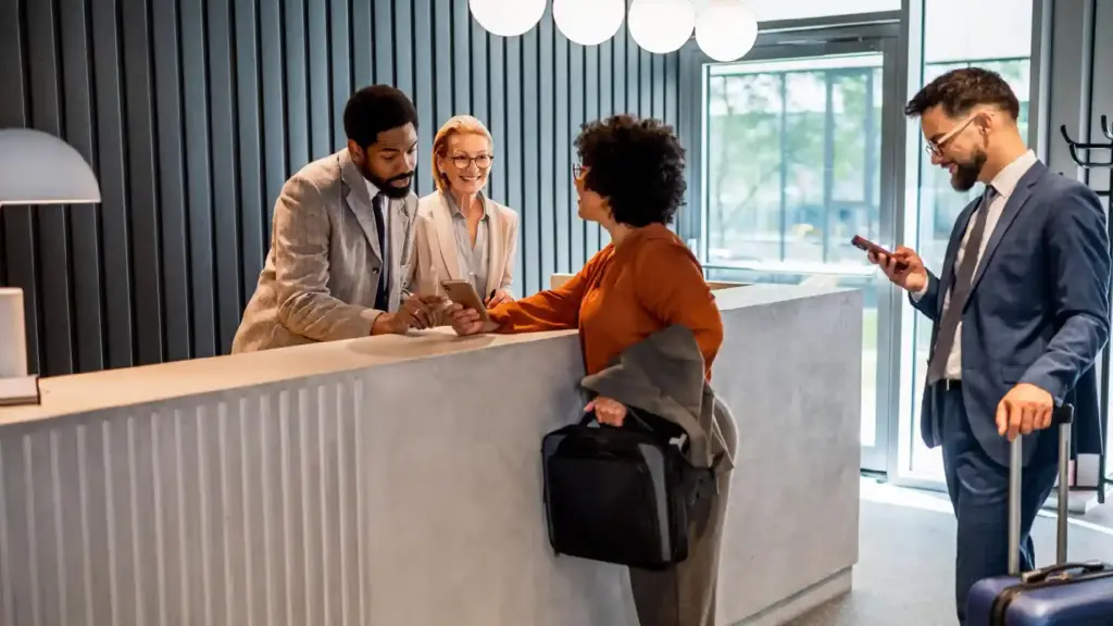 Four professionals check in at a hotel reception, talking with a smiling woman while another man checks his phone with a suitcase nearby.