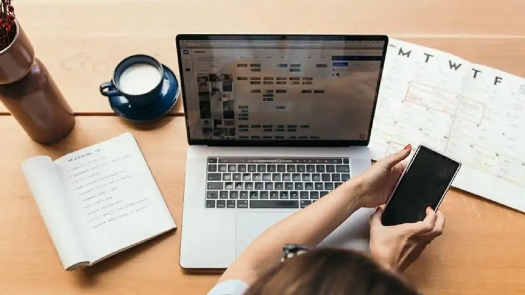 Overhead view of a workspace with a laptop, smartphone, open notebook, cup of coffee, and a calendar on a wooden desk.