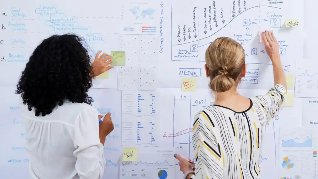 Two people stand with their backs to the camera, writing on a wall covered with charts, graphs, and sticky notes during a brainstorming session.