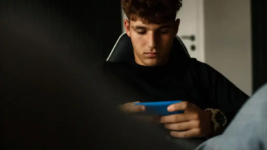 Young man in a black shirt sits in a dim room, looking down at a blue handheld device in his hands.