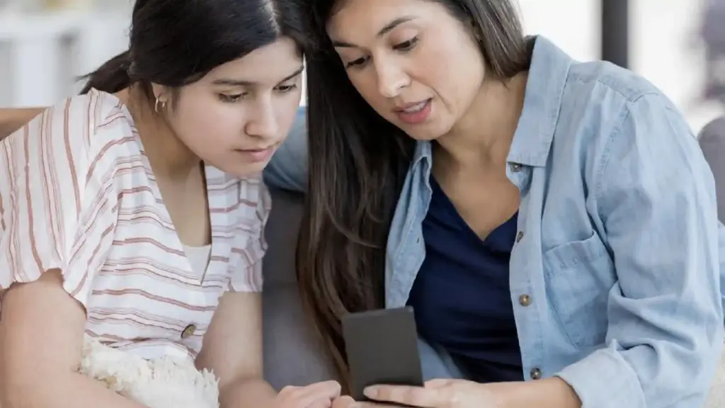 Two women sit close together on a couch, looking at a smartphone held between them.
