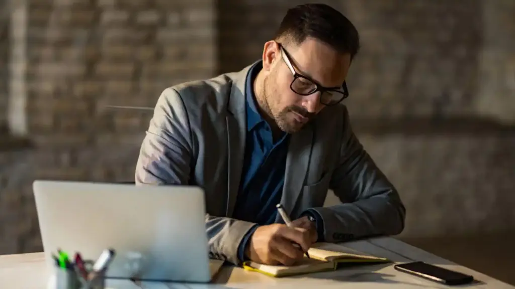 New Project 86 - Brill Creations Man in a blazer and glasses writing in a notebook at a desk with an open laptop beside him