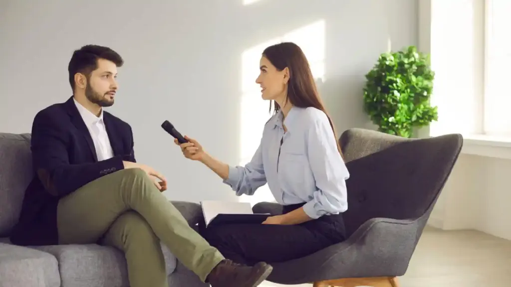 Woman interviewer holds a microphone to a man seated on a gray sofa for an interview in a bright lounge area.