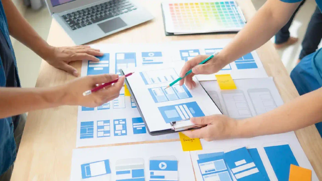 Team members collaborate over blue UI wireframes and sticky notes on a table, with a laptop nearby. (design planning)