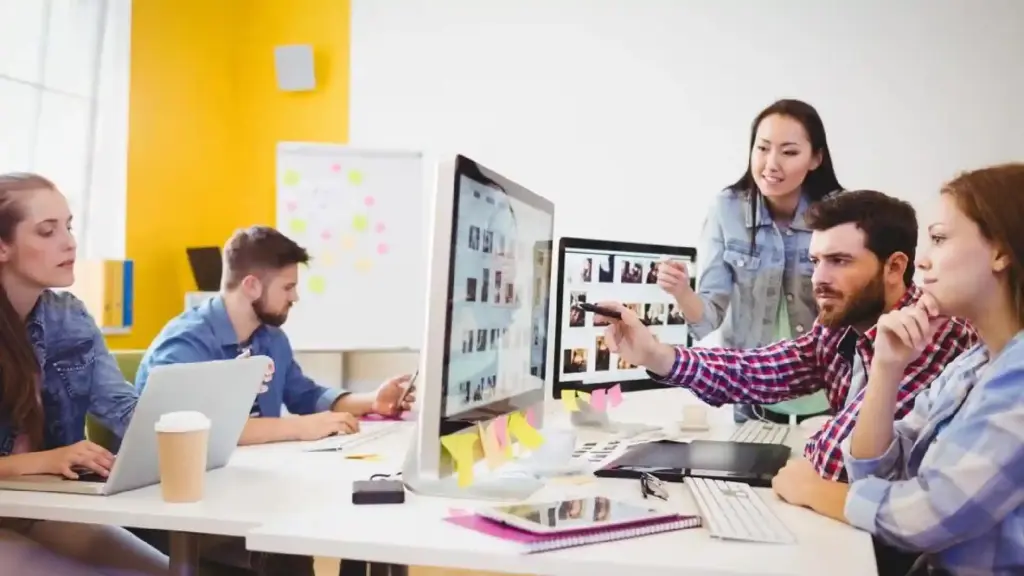 Team of coworkers collaborating at a desk, looking at two large monitors with colorful sticky notes nearby